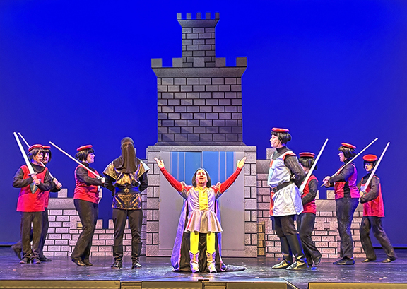A photo of a musical number from the Shrek play. Lord Farquaad, a short guy, stands at the center with his arms raised. He is flanked by his royal guards on either side.